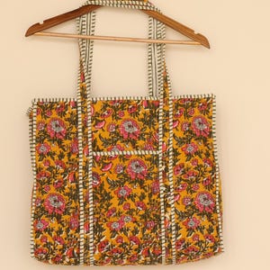 Op de afbeelding: Een gele tote bag met een bloemenpatroon in roze en groen. De tas heeft een rechthoekige vorm met een zak aan de voorkant en wit en zwart gestreepte rand. De tas hangt aan een houten hanger.