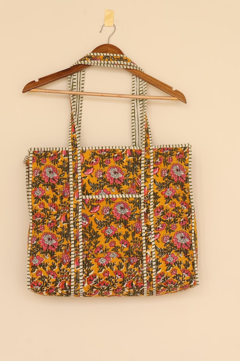 Op de afbeelding: Een gele tote bag met een bloemenpatroon in roze en groen. De tas heeft een rechthoekige vorm met een zak aan de voorkant en wit en zwart gestreepte rand. De tas hangt aan een houten hanger.