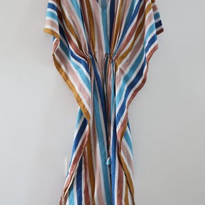 May include: A striped caftan in shades of blue, brown, pink, and gold hangs on a wooden hanger. The caftan features a V-neck and a drawstring waist. The fabric has vertical stripes.