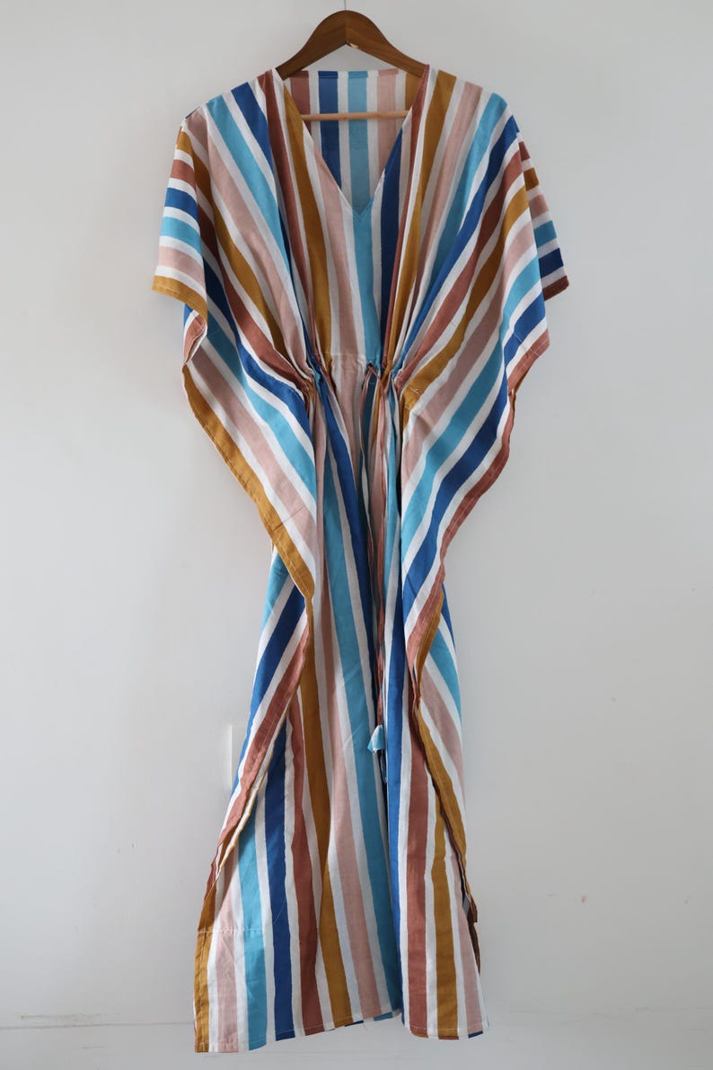 May include: A striped caftan in shades of blue, brown, pink, and gold hangs on a wooden hanger. The caftan features a V-neck and a drawstring waist. The fabric has vertical stripes.