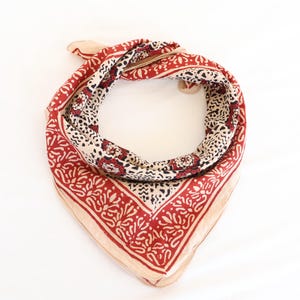 May include: A square bandana with a repeating floral and geometric pattern in red, cream, and black. The bandana is folded and tied, showcasing the detailed design. The fabric appears to be lightweight.