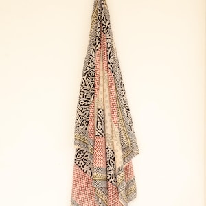 May include: A patterned scarf with a variety of designs, including paisley and zigzags, hangs against a white background. The scarf features a color palette of black, red, beige, and olive green. The scarf is made of a lightweight fabric.