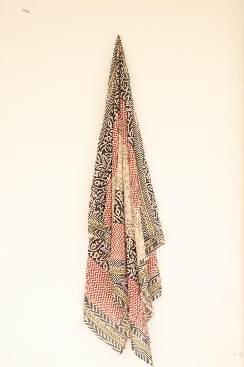 May include: A patterned scarf with a variety of designs, including paisley and zigzags, hangs against a white background. The scarf features a color palette of black, red, beige, and olive green. The scarf is made of a lightweight fabric.