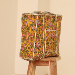 Op de afbeelding: Een grote tote bag met een bloemenpatroon in geel, roze en groen. De tas heeft een rechthoekige vorm met handvatten en een wit en zwart gestikte rand. Hij staat op een kleine houten kruk.