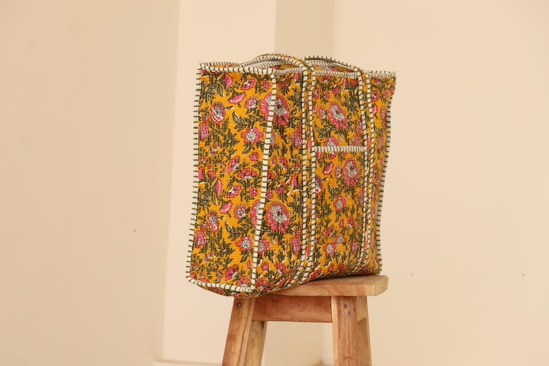 Op de afbeelding: Een grote tote bag met een bloemenpatroon in geel, roze en groen. De tas heeft een rechthoekige vorm met handvatten en een wit en zwart gestikte rand. Hij staat op een kleine houten kruk.