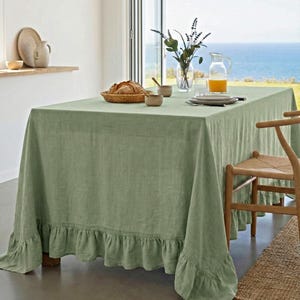 Puede incluir: Un mantel de lino verde salvia con borde con volantes cubre una mesa de madera. La mesa está puesta con una cesta de pan tejida, cuencos pequeños, platos y una jarra de cristal con zumo de naranja. Una silla de madera está al lado de la mesa.