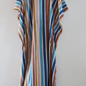 May include: A colorful, striped caftan dress in shades of blue, brown, pink, and white. The dress is hanging on a wooden hanger against a white background. The vertical stripes create a flowing, relaxed silhouette, perfect for casual wear.