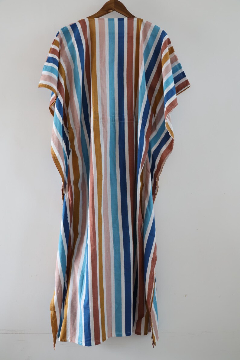 May include: A colorful, striped caftan dress in shades of blue, brown, pink, and white. The dress is hanging on a wooden hanger against a white background. The vertical stripes create a flowing, relaxed silhouette, perfect for casual wear.