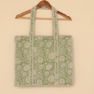 Pu&ograve; includere: Una borsa tote con stampa floreale verde e bianca con manici abbinati, appesa a una gruccia di legno. La borsa presenta un motivo ripetuto di fiori e foglie, con un bordo e cuciture bianche.