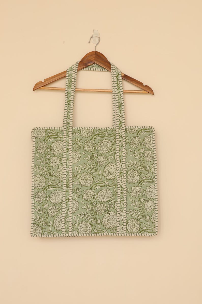 Pu&ograve; includere: Una borsa tote con stampa floreale verde e bianca con manici abbinati, appesa a una gruccia di legno. La borsa presenta un motivo ripetuto di fiori e foglie, con un bordo e cuciture bianche.