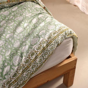 May include: A green and white floral patterned duvet cover and pillowcase set on a wooden bed frame. The bedding features a repeating floral design with a border of paisley and geometric patterns. The color palette includes shades of green, white, and gold.
