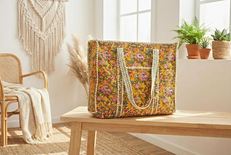 Op de afbeelding: Een grote, rechthoekige draagtas met een bloemenpatroon in geel, roze en groen staat op een houten bank. De tas heeft cr&egrave;mekleurige handvatten en randen. De achtergrond omvat een macram&eacute; wandkleed, een rieten stoel en potplanten.