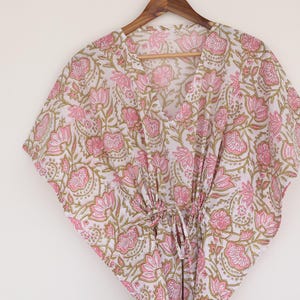 May include: A white and pink floral print kaftan dress hanging on a wooden hanger. The dress features a V-neckline and a drawstring waist. The floral pattern includes pink flowers and green leaves.