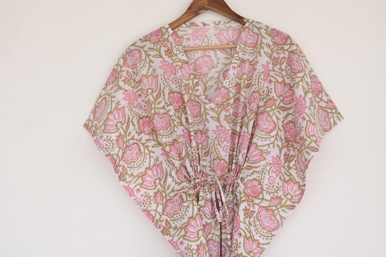 May include: A white and pink floral print kaftan dress hanging on a wooden hanger. The dress features a V-neckline and a drawstring waist. The floral pattern includes pink flowers and green leaves.