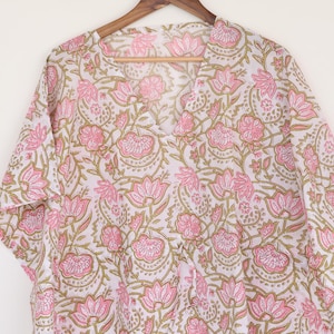 May include: A white and pink floral print tunic top with a V-neck and short sleeves. The fabric features a repeating pattern of pink flowers and green leaves. The top is hanging on a wooden hanger.