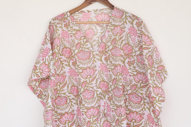May include: A white and pink floral print tunic top with a V-neck and short sleeves. The fabric features a repeating pattern of pink flowers and green leaves. The top is hanging on a wooden hanger.