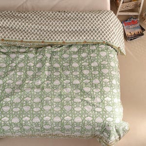 May include: A green and white floral patterned quilt on a bed. The quilt features a repeating pattern of white flowers on a green background. The top of the quilt has a smaller, repeating pattern in green and white. The quilt is folded over a white sheet.