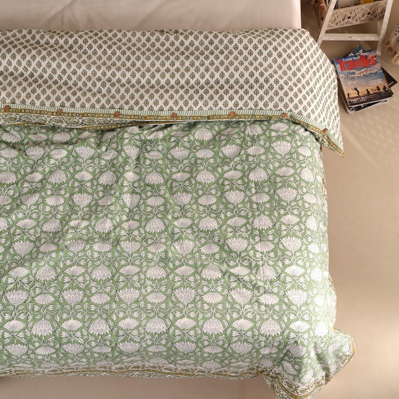 May include: A green and white floral patterned quilt on a bed. The quilt features a repeating pattern of white flowers on a green background. The top of the quilt has a smaller, repeating pattern in green and white. The quilt is folded over a white sheet.