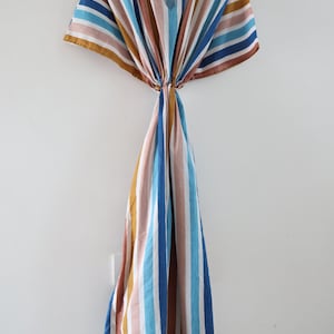 May include: A striped caftan dress in a variety of colors, including blue, brown, pink, and white, hangs on a wooden hanger. The dress features a V-neck and a flowing, loose fit, perfect for warm weather. The vertical stripes create a visually appealing design.