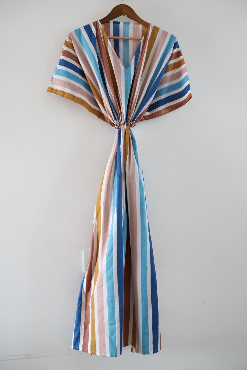May include: A striped caftan dress in a variety of colors, including blue, brown, pink, and white, hangs on a wooden hanger. The dress features a V-neck and a flowing, loose fit, perfect for warm weather. The vertical stripes create a visually appealing design.