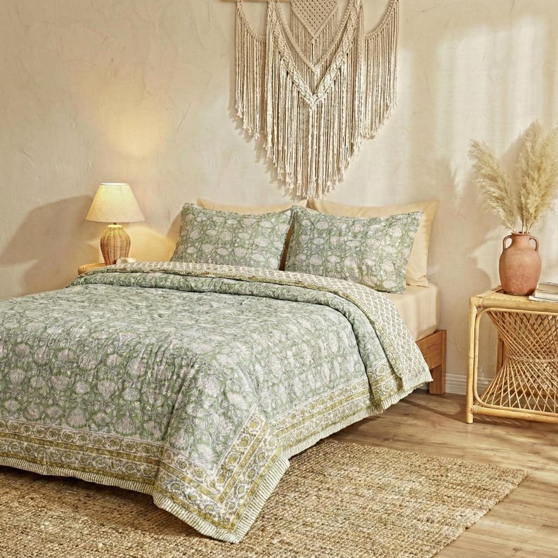 May include: A bed with a green and white floral patterned quilt and matching pillowcases. The quilt has a decorative border with a yellow and green design. A woven wall hanging and a small table with a lamp are visible.