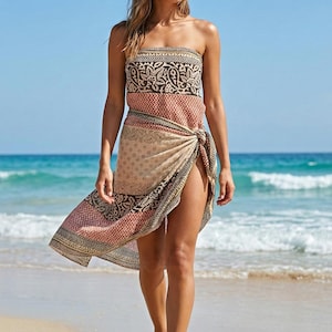 May include: A woman wearing a straw hat and a patterned sarong dress on a beach. The dress features a mix of brown, beige, and red patterns. The ocean and blue sky are in the background.