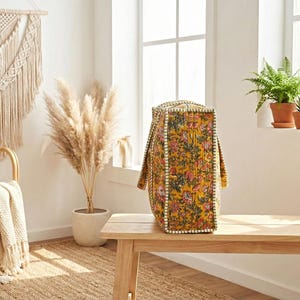 Op de afbeelding: Een gele tote bag met een bloemenpatroon, geplaatst op een houten bank. De tas heeft een rechthoekige vorm met handvatten en een witte kralenrand. De achtergrond omvat een macram&eacute; wandkleed en potplanten.