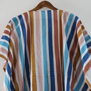 May include: A colorful, striped garment hangs on a wooden hanger. The fabric features vertical stripes in shades of blue, brown, yellow, and pink. The garment has short sleeves and a loose fit.