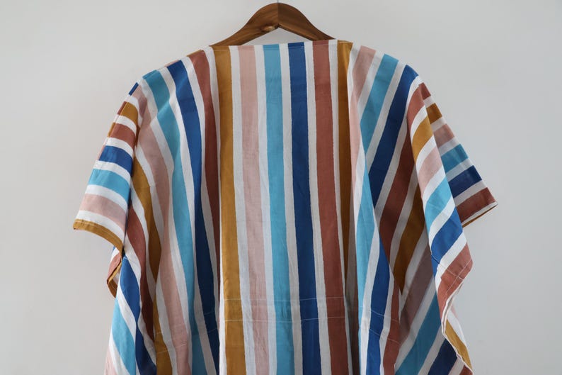 May include: A colorful, striped garment hangs on a wooden hanger. The fabric features vertical stripes in shades of blue, brown, yellow, and pink. The garment has short sleeves and a loose fit.