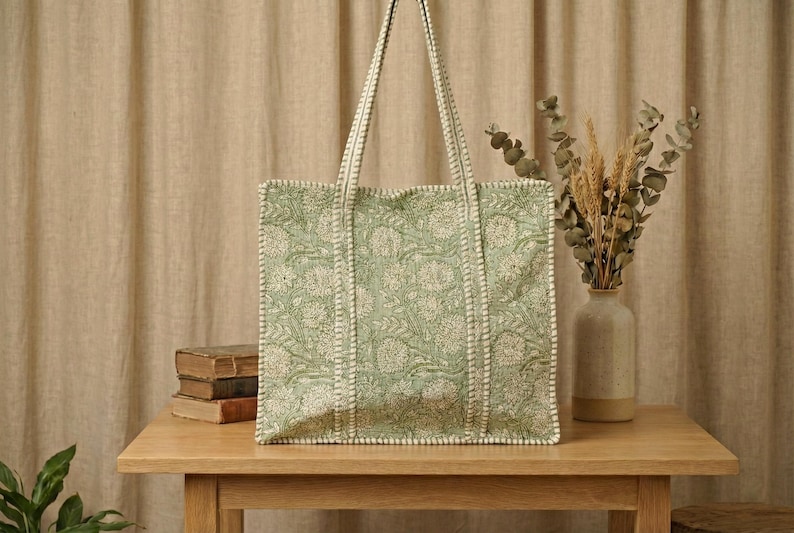 Pu&ograve; includere: Una borsa tote verde chiaro con un motivo floreale e bordi bianchi. La borsa ha manici lunghi ed &egrave; appoggiata su un tavolo di legno. Un vaso con piante secche &egrave; a destra della borsa.
