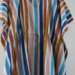 May include: A colorful, striped caftan dress in a variety of vertical stripes. The stripes include shades of blue, brown, white, and yellow. The dress has a V-neck and a drawstring waist. The caftan is hanging on a wooden hanger.