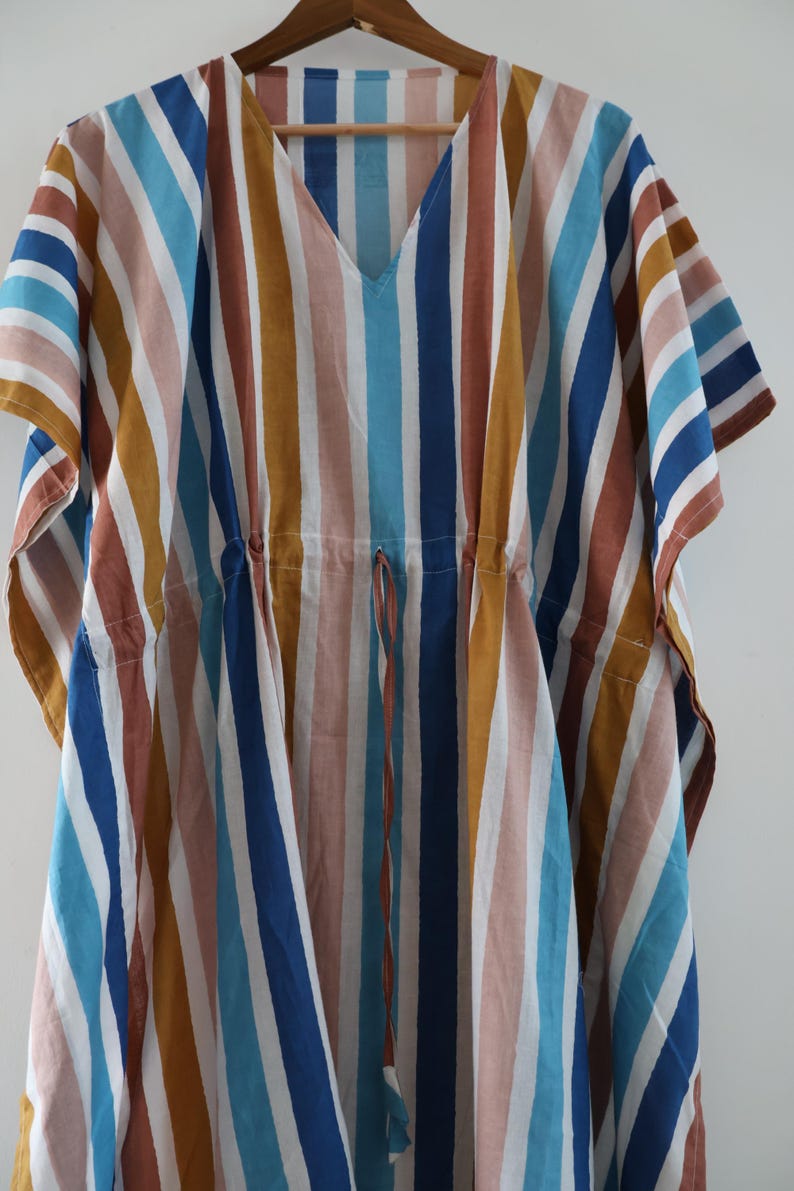 May include: A colorful, striped caftan dress in a variety of vertical stripes. The stripes include shades of blue, brown, white, and yellow. The dress has a V-neck and a drawstring waist. The caftan is hanging on a wooden hanger.