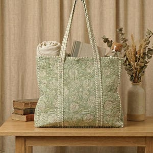 Pu&ograve; includere: Una grande borsa tote verde con un motivo floreale, riempita con una coperta arrotolata, libri e altri oggetti. La borsa &egrave; su un tavolo di legno, con un vaso di fiori secchi e un barattolo con coperchio di legno.