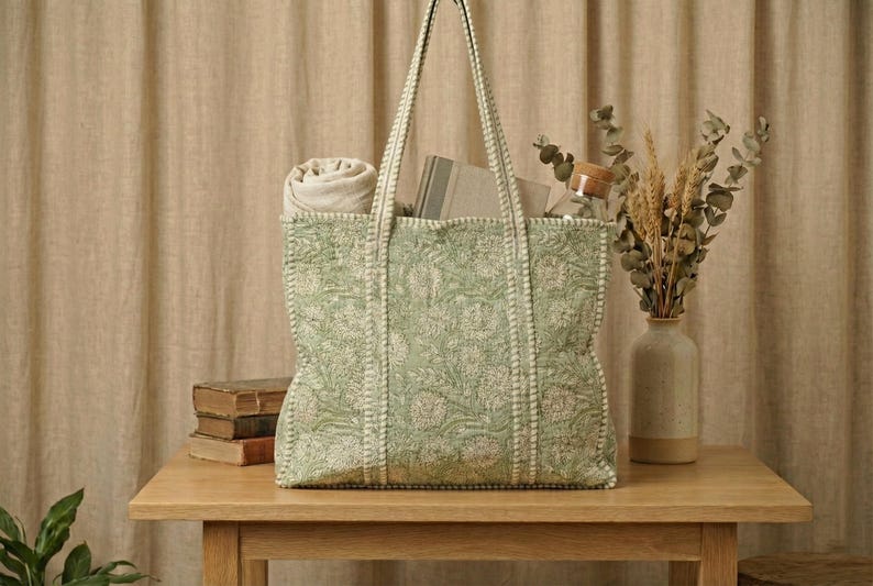Pu&ograve; includere: Una grande borsa tote verde con un motivo floreale, riempita con una coperta arrotolata, libri e altri oggetti. La borsa &egrave; su un tavolo di legno, con un vaso di fiori secchi e un barattolo con coperchio di legno.