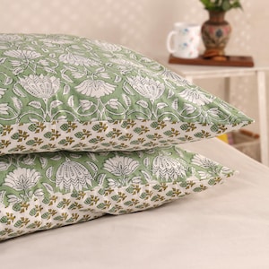 May include: Two green and white floral patterned pillowcases stacked on top of each other. The pillowcases have a repeating pattern of large white flowers and leaves on a light green background. The bottom edge features a border of small brown and white floral designs.