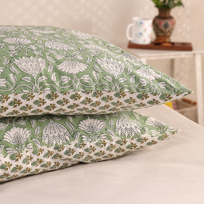 May include: Two green and white floral patterned pillowcases stacked on top of each other. The pillowcases have a repeating pattern of large white flowers and leaves on a light green background. The bottom edge features a border of small brown and white floral designs.