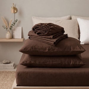 May include: A set of dark brown bedding featuring a fitted sheet, a flat sheet, and two pillowcases. The fabric has a visible, textured weave. The items are stacked neatly on a bed, showcasing the deep, solid brown color of the textile material.