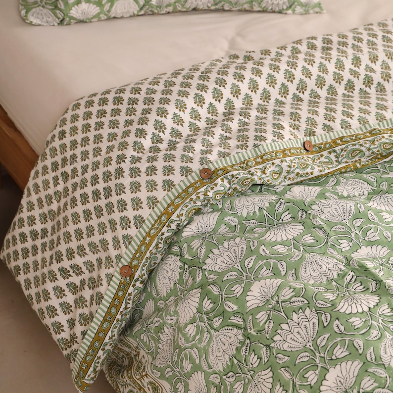 May include: A green and white floral patterned quilt with a matching pillow. The quilt features a repeating pattern of small green floral motifs on a white background, and a larger floral design on the bottom. Wooden buttons secure the quilt.