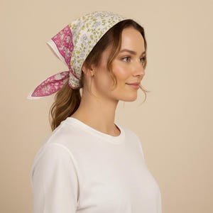 May include: A floral print headscarf in pink, white, and green is tied around the head. The scarf is worn with a white crew neck shirt. The background is a solid beige color.