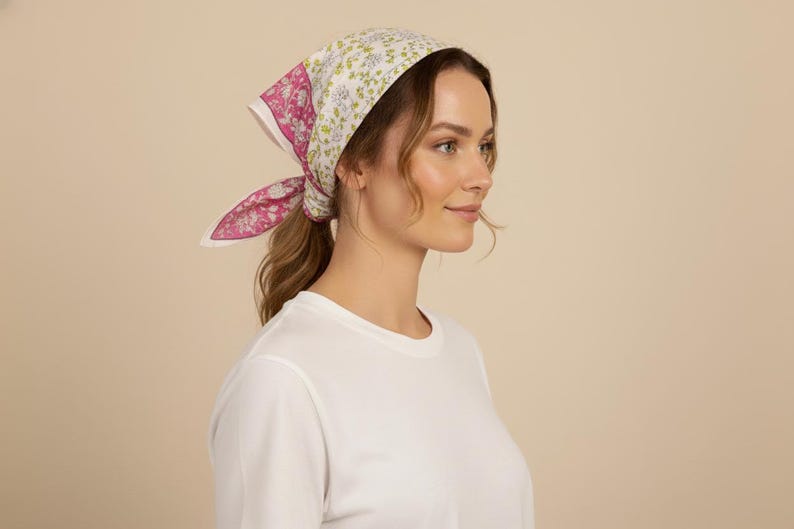 May include: A floral print headscarf in pink, white, and green is tied around the head. The scarf is worn with a white crew neck shirt. The background is a solid beige color.