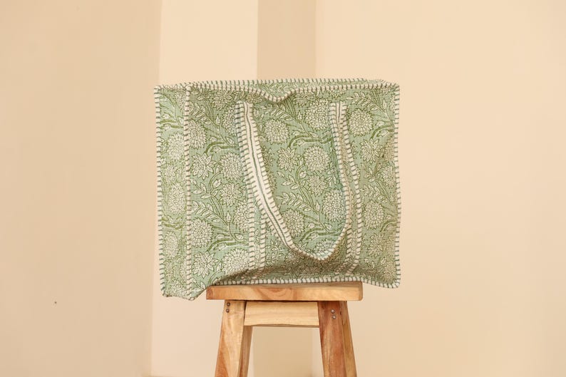Pu&ograve; includere: Una grande borsa tote quadrata con un motivo floreale nei toni del verde e del bianco. La borsa ha due manici ed &egrave; appoggiata su uno sgabello di legno. Il design della borsa presenta una stampa floreale ripetuta.