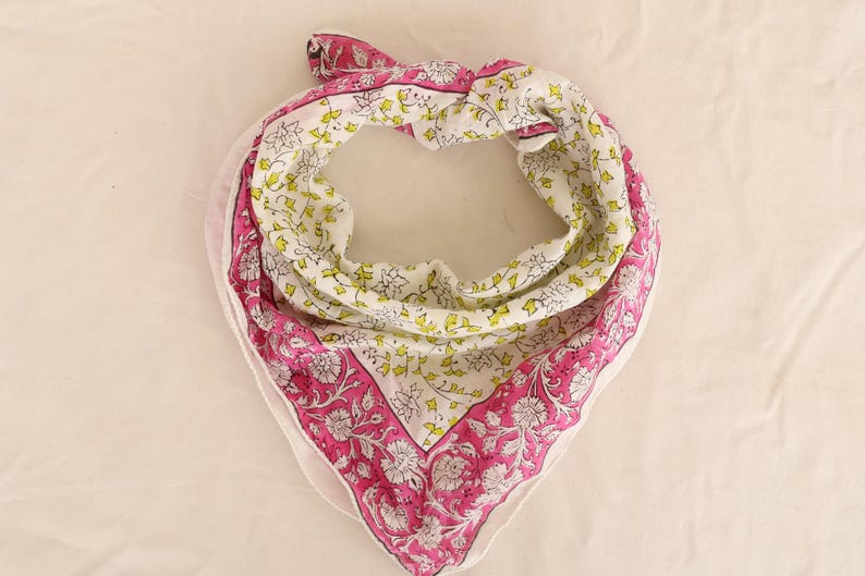 May include: A square scarf with a floral pattern. The scarf features a white background with a repeating pattern of yellow and green leaves. The border is a vibrant pink with a floral design. The scarf is folded and tied.