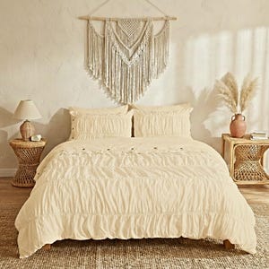 May include: A cream-coloured bed with matching pillows and a duvet. The bed is made up with a textured fabric. A woven wall hanging hangs above the bed. A wicker side table with a lamp and a vase with dried flowers are on either side of the bed.