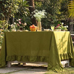 Puede incluir: Una mesa de jardín cubierta con un mantel verde con un borde con volantes. La mesa está puesta con platos, vasos y un cuenco de naranjas. Un jarrón con flores blancas y otras plantas se encuentran en el fondo.