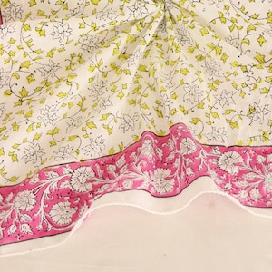 May include: A white textile with a floral pattern and a pink border. The border features a repeating floral design. The center of the textile has a pattern of white and yellow flowers and leaves.