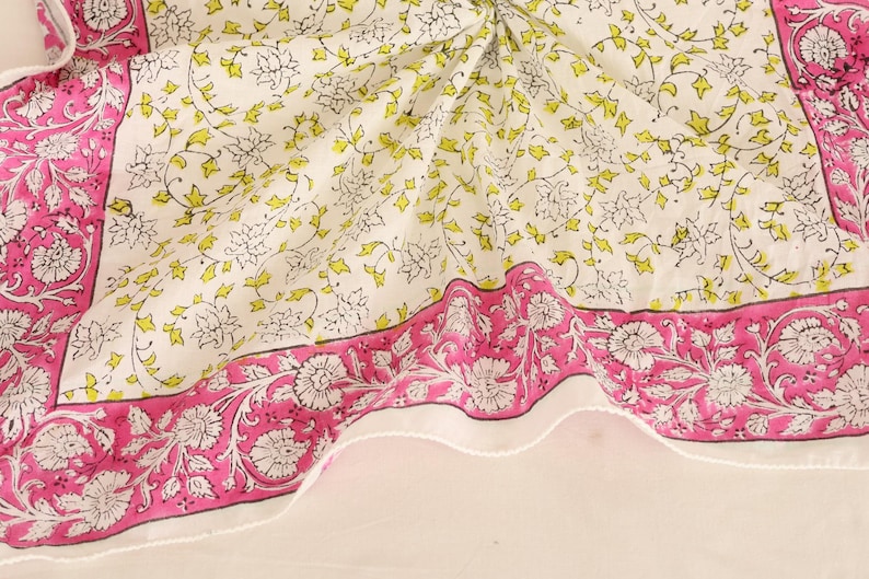 May include: A white textile with a floral pattern and a pink border. The border features a repeating floral design. The center of the textile has a pattern of white and yellow flowers and leaves.