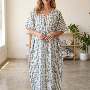 May include: A woman wearing a white and teal patterned caftan dress with a V-neck and drawstring waist. The dress features a repeating fish design. The model is wearing brown sandals. The background includes a wooden shelf with rolled towels and potted plants.