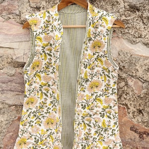 Può includere: Un gilet senza maniche con apertura frontale e motivo floreale. Il gilet presenta uno sfondo bianco con fiori gialli e pesca e foglie verdi. La fodera interna ha strisce verticali verdi e bianche.