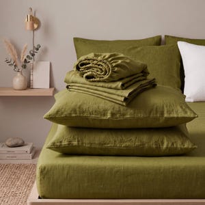 Peut inclure: Un ensemble de linge de lit vert olive comprenant un drap-housse, un drap plat et deux taies d'oreiller. Le tissu présente une texture tissée visible. Les draps sont soigneusement pliés et empilés sur le lit, créant un aspect superposé.