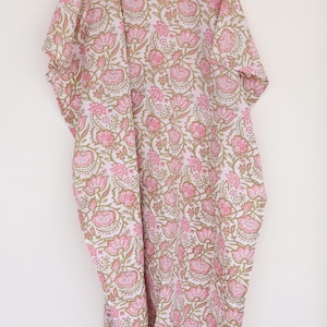 May include: A white and pink floral print kaftan dress hanging on a wooden hanger. The dress features a repeating pattern of pink flowers and green leaves. The kaftan is made of a lightweight fabric.